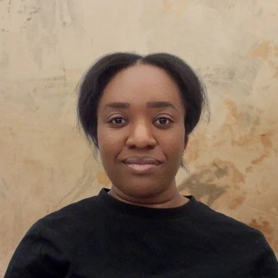A woman with short black hair wearing a black top, standing in front of a beige and brown textured wall.
