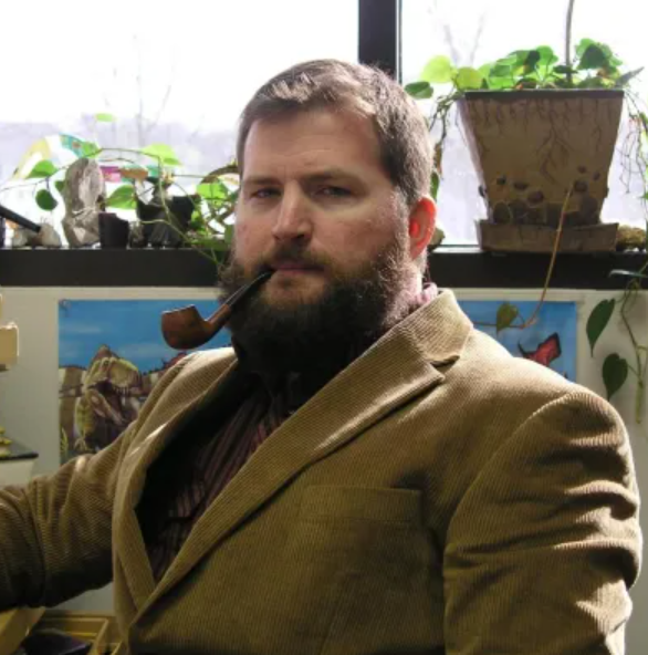 Man with beard smoking a pipe in an office, with plants and windows in the background.
