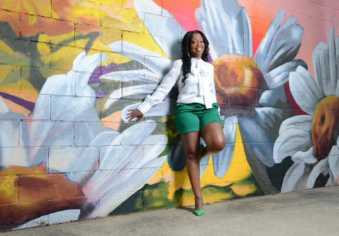 A woman in a white blouse and green shorts is posing against a colorful floral mural painted on a brick wall.
