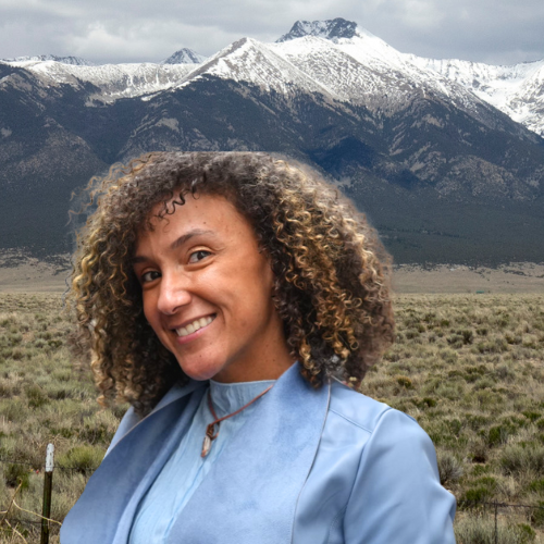 Close-up of a smiling Stephanie Bowman Campaigns & Trainings Manager, a Black woman with curly hair smiling outdoors in front of snow-capped mountains and a grassy plain.