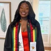 A woman smiling, wearing a graduation gown and a colorful sash, standing indoors with framed artwork and window blinds behind her.