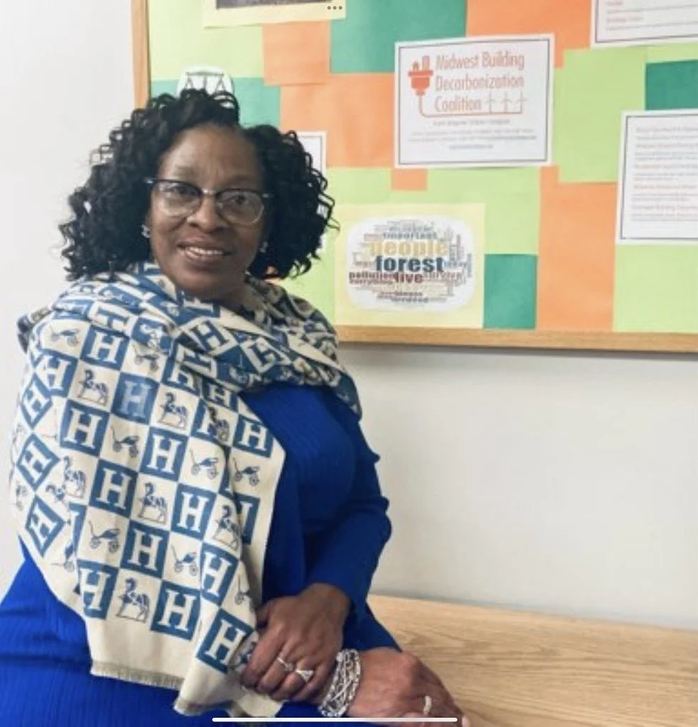 A woman with glasses and curly hair wearing a blue dress and a white scarf with blue 'H' logos, standing in front of a colorful bulletin board.
