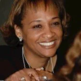 A woman smiling and wearing earrings, possibly at a social event.