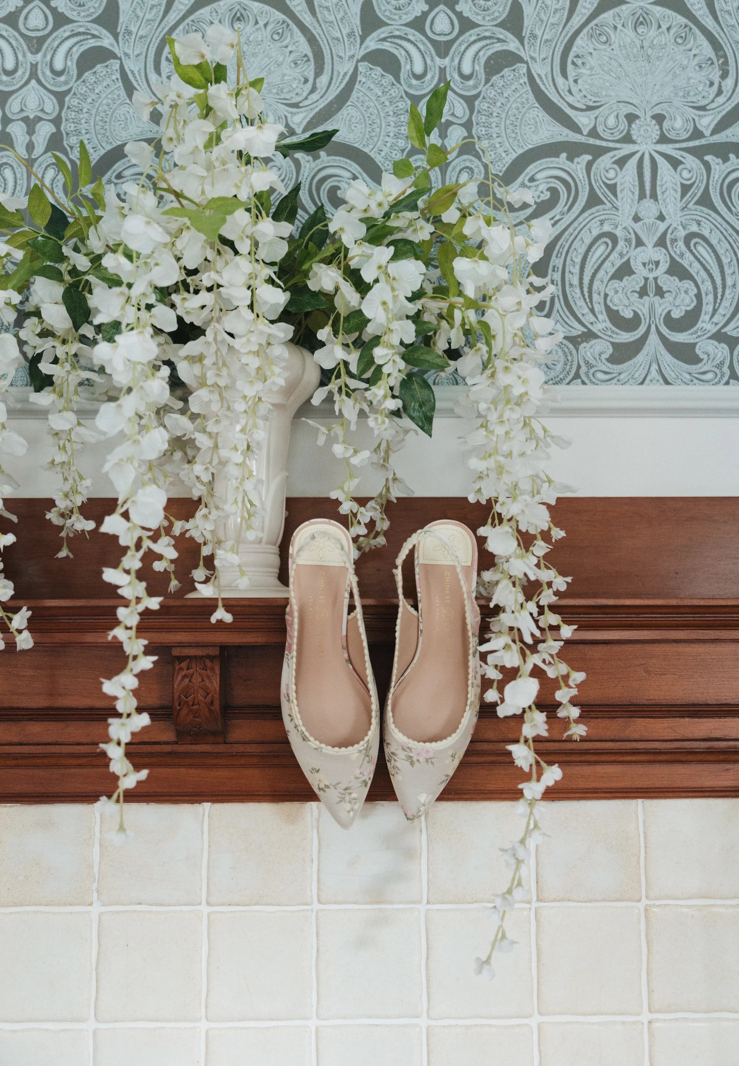 A pair of elegant white high-heeled shoes with floral embroidery placed on a wooden mantel beneath a cascading bouquet of white flowers in a white vase, against a blue and white ornate wallpaper background.