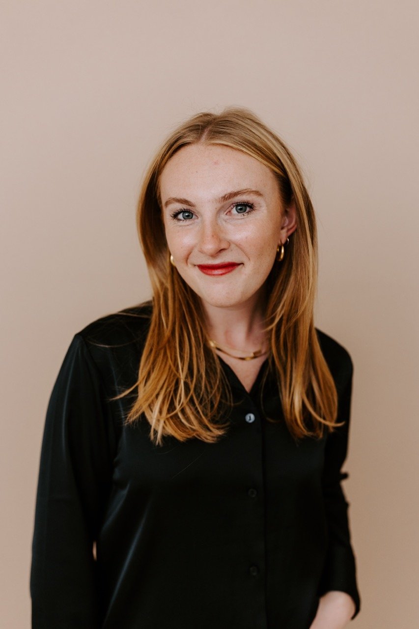 A young woman with red hair, blue eyes, and light skin, wearing a black button-up shirt, gold jewelry, and red lipstick, smiling against a plain beige background.