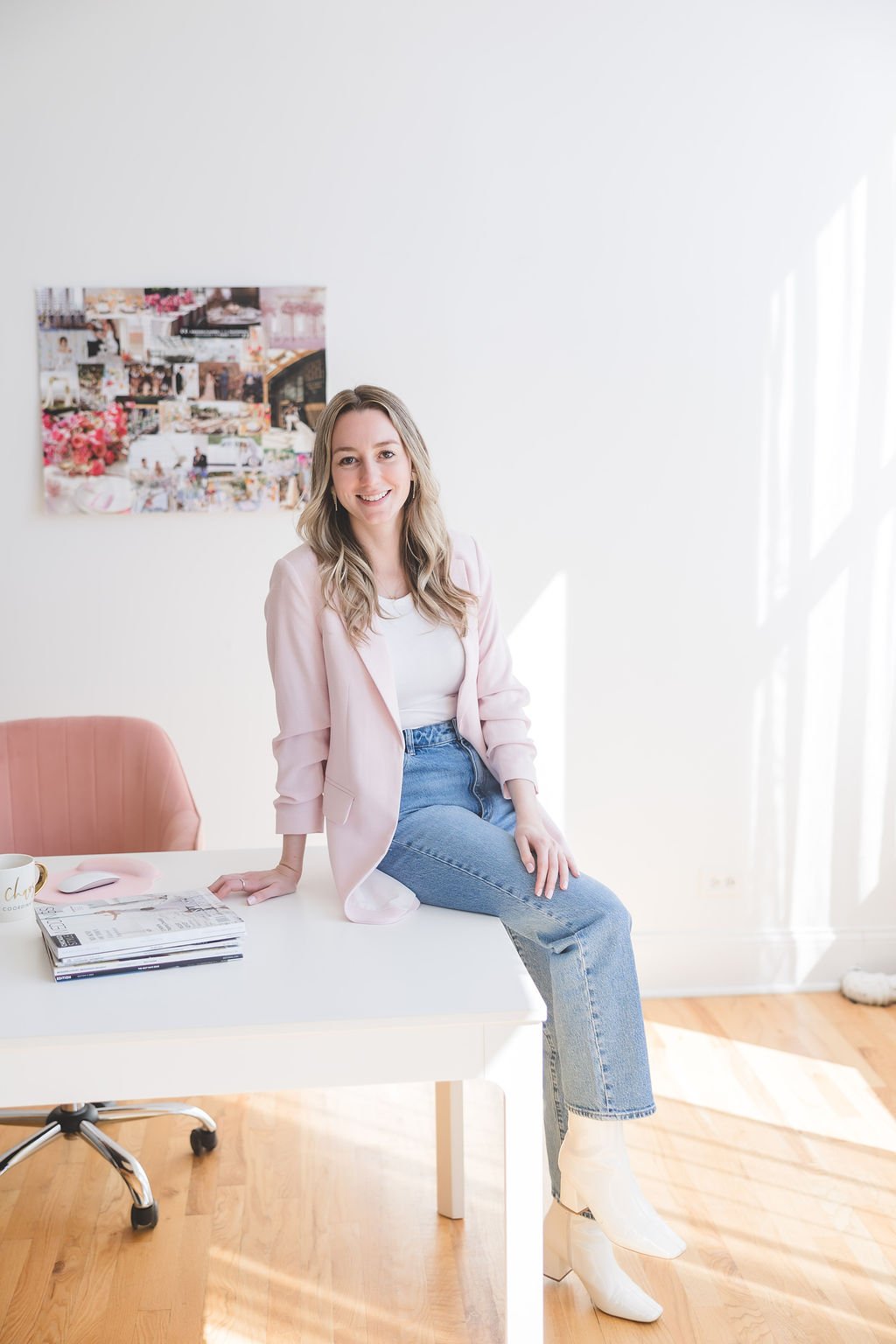 A young woman with blonde hair, wearing a pink blazer, white shirt, and jeans, sitting on the edge of a white desk in a bright, minimalistic office space. She is smiling and looking at the camera. There is a pink chair, a stack of magazines, and a coffee mug on the desk. A collage of photos hangs on the plain white wall behind her, and sunlight streams through a window on the right side.