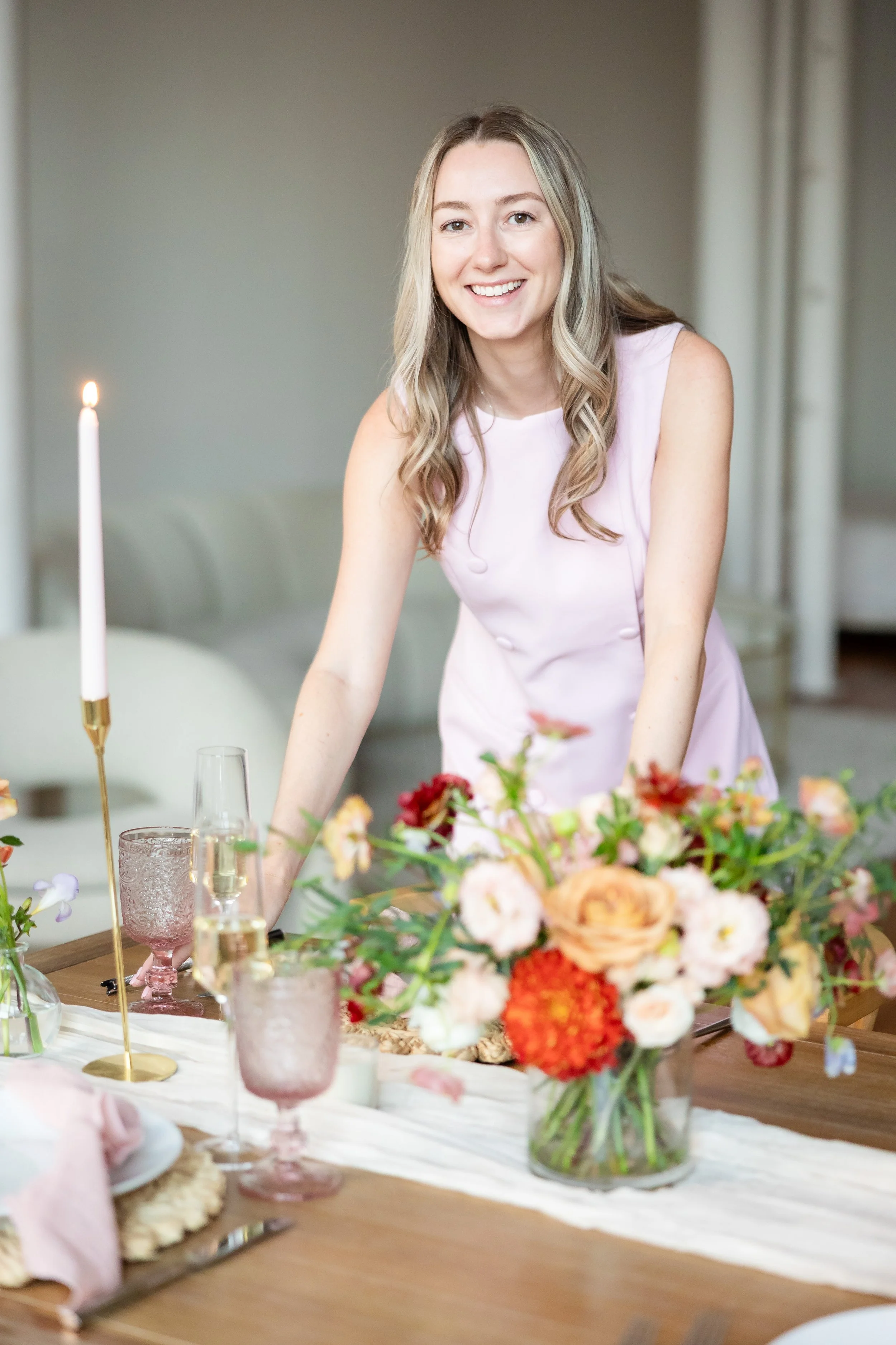 A woman with long blonde hair wearing a light pink dress, smiling at the camera, standing behind a dining table decorated with a floral arrangement, glasses, and candles.