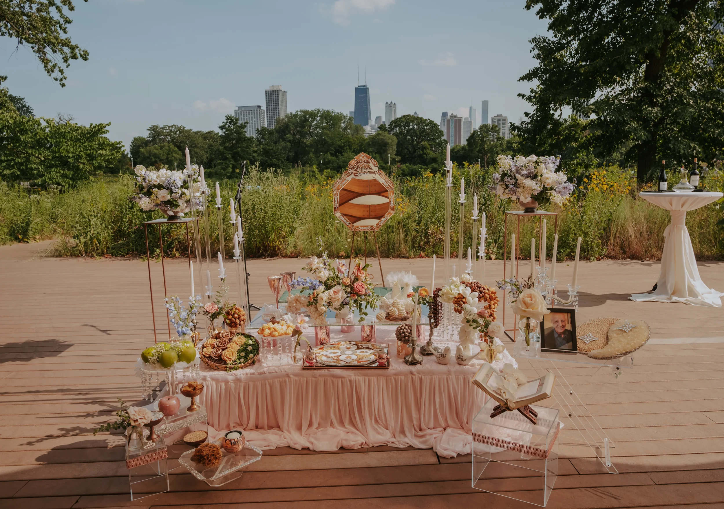 Elegant outdoor table setup with pink draped tablecloth, floral arrangements, candles, decorative items, and city skyline in the background.