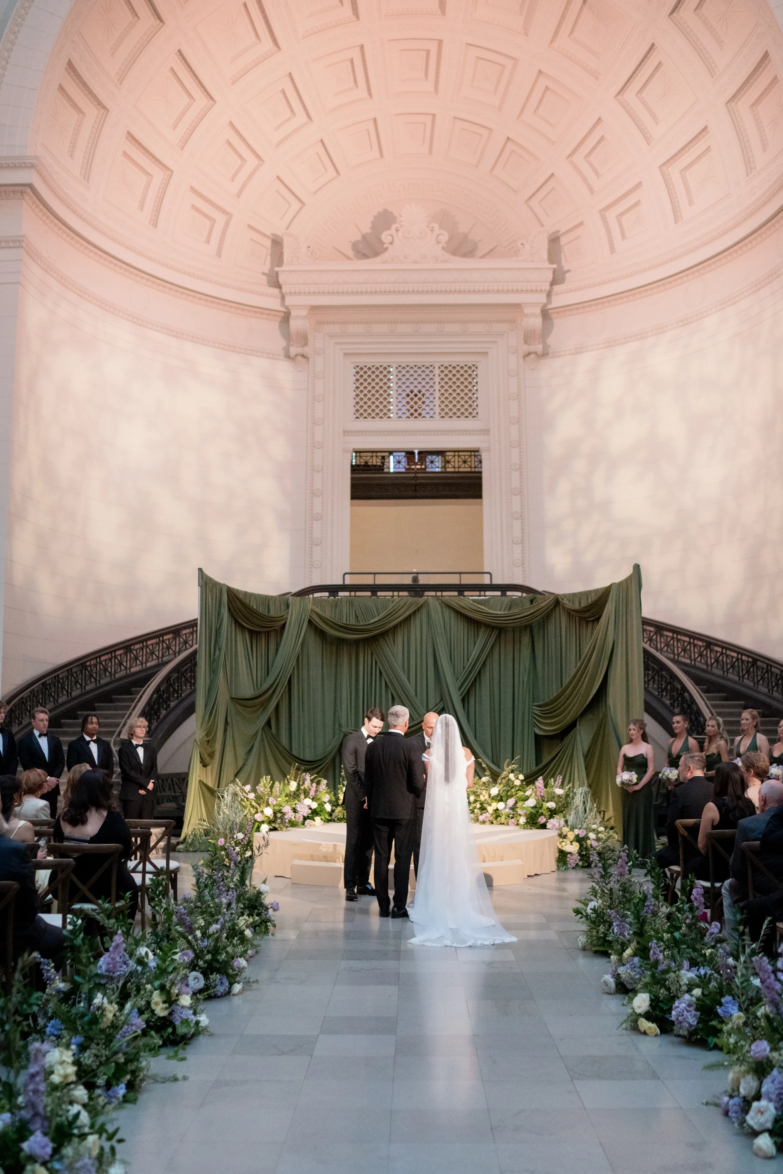 A wedding ceremony taking place inside a large, ornate building with a high, decorated ceiling and elegant architectural details. The couple is standing under a green draped backdrop with flowers, while guests and attendants watch. Afterglow Weddings