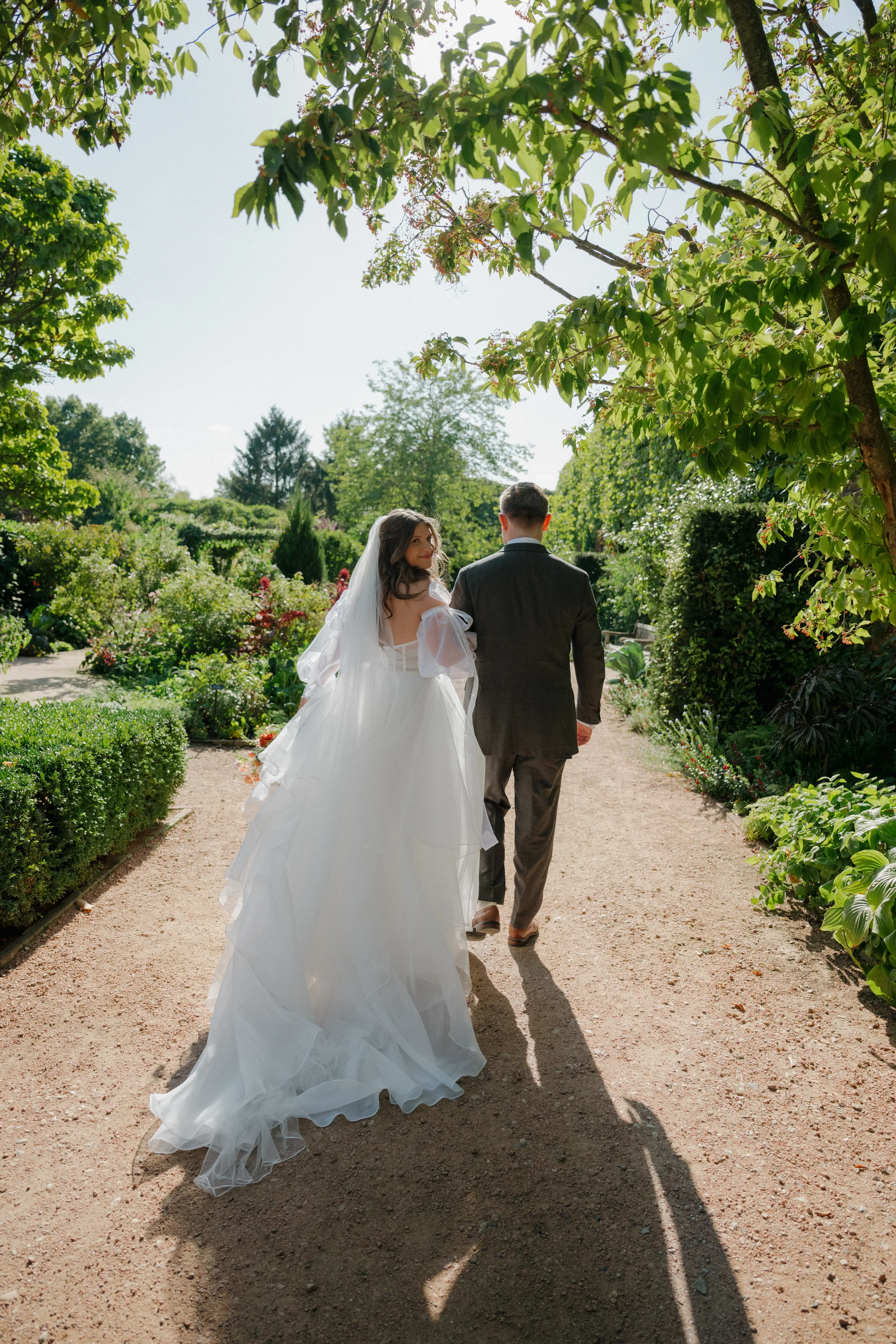 Bride and groom walking down a garden path on their wedding day, surrounded by lush green trees and shrubs, with sunlight shining through the leaves.
