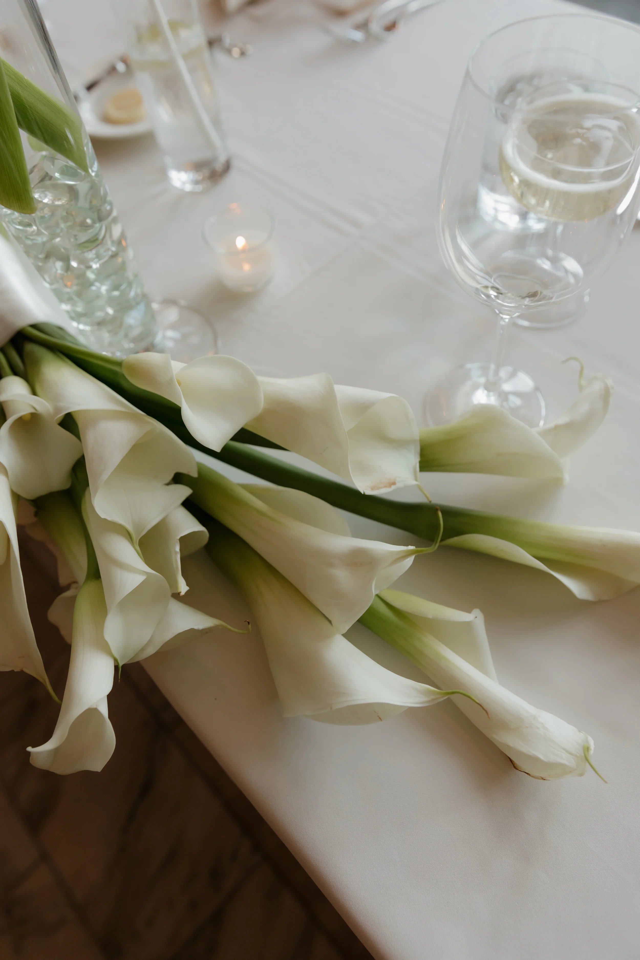 White calla lilies on a table with a glass of water and a water pitcher, candles, and a white tablecloth.