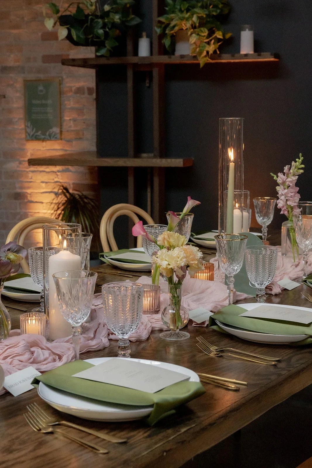 Elegant dining table set with candles, flowers, glassware, and plates, in a warmly lit room with brick wall and tope shelves with plants, showcased from a wedding by Afterglow, Chicago Wedding Planners & Coordinators.