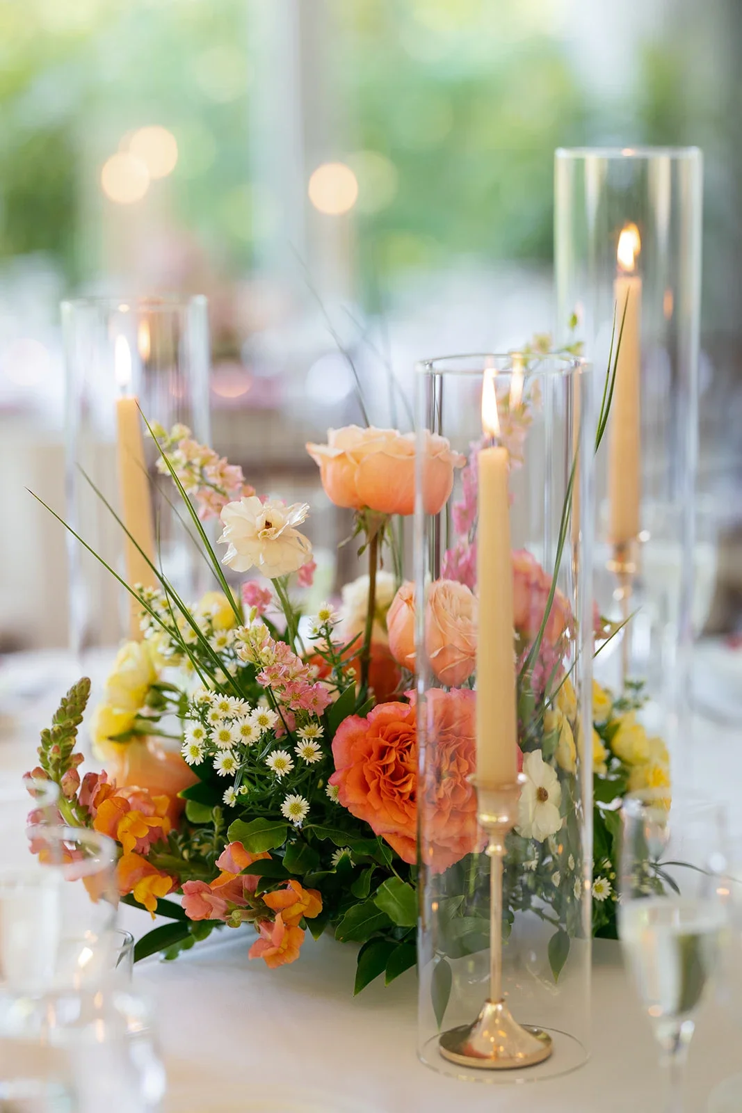 Elegant floral centerpiece with pink, yellow, and white flowers in a glass vase, surrounded by tall candles in glass holders, on a white table with blurred background, showcased from a wedding by Afterglow, Chicago Wedding Planners & Coordinators.