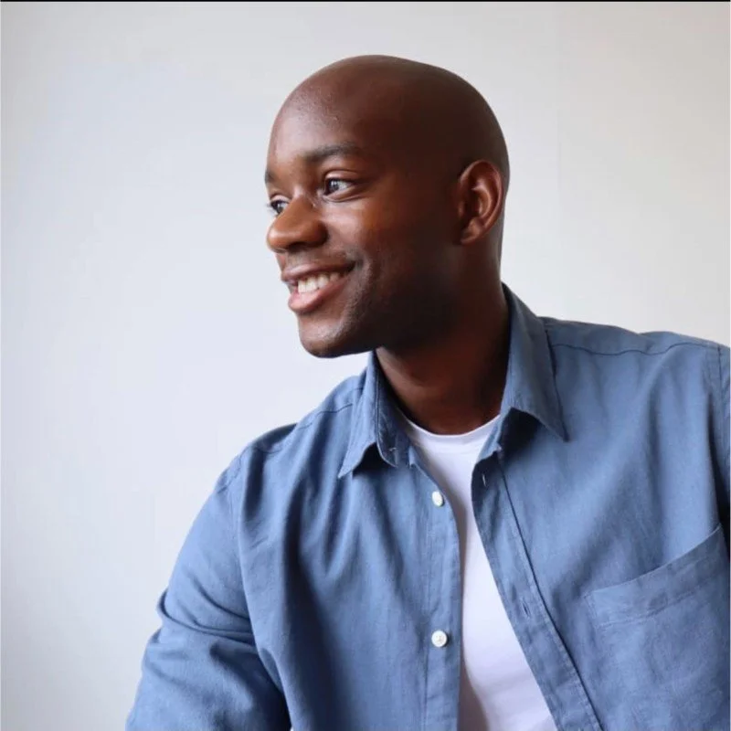 A smiling Black man with a shaved head wearing a denim shirt and white undershirt, looking to the side against a plain light background.