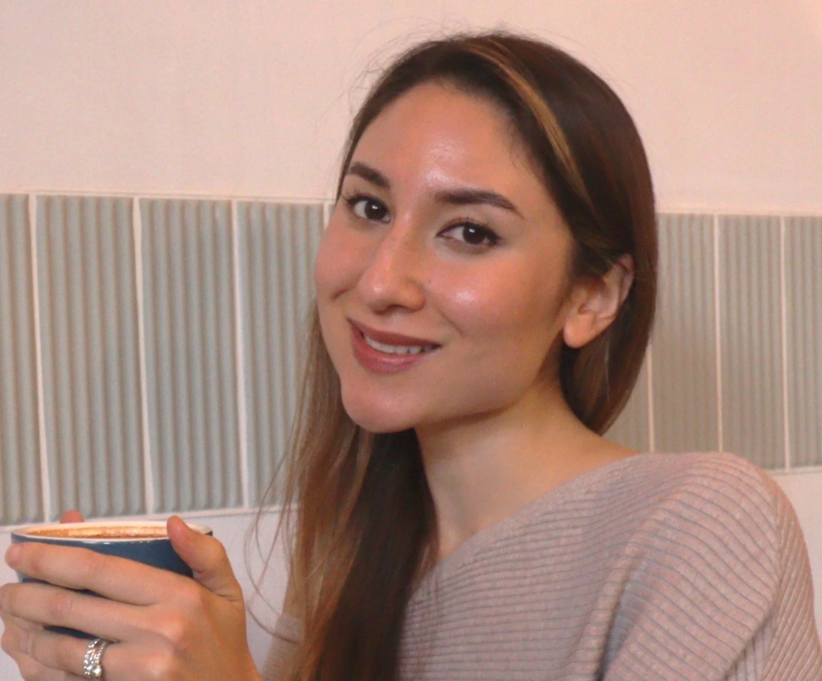 A smiling woman with light skin and long brown hair holding a dark blue coffee cup, standing in front of beige and white tiled wall.