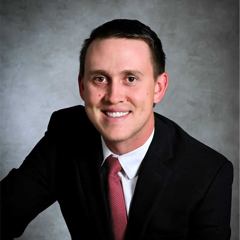Headshot of a smiling man in a black suit, white shirt, and red tie, against a gray background.