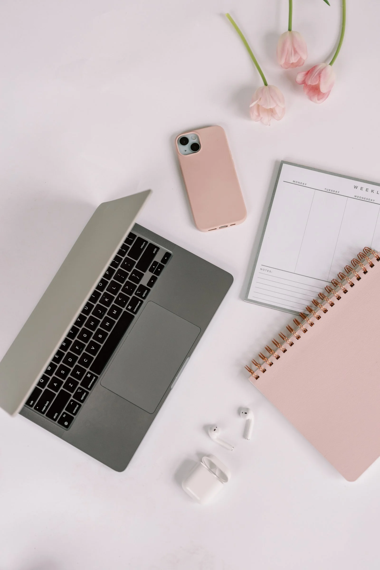 Open laptop, smartphone with pink case, weekly planner, pink notepad, AirPods, and pink tulips on white background.