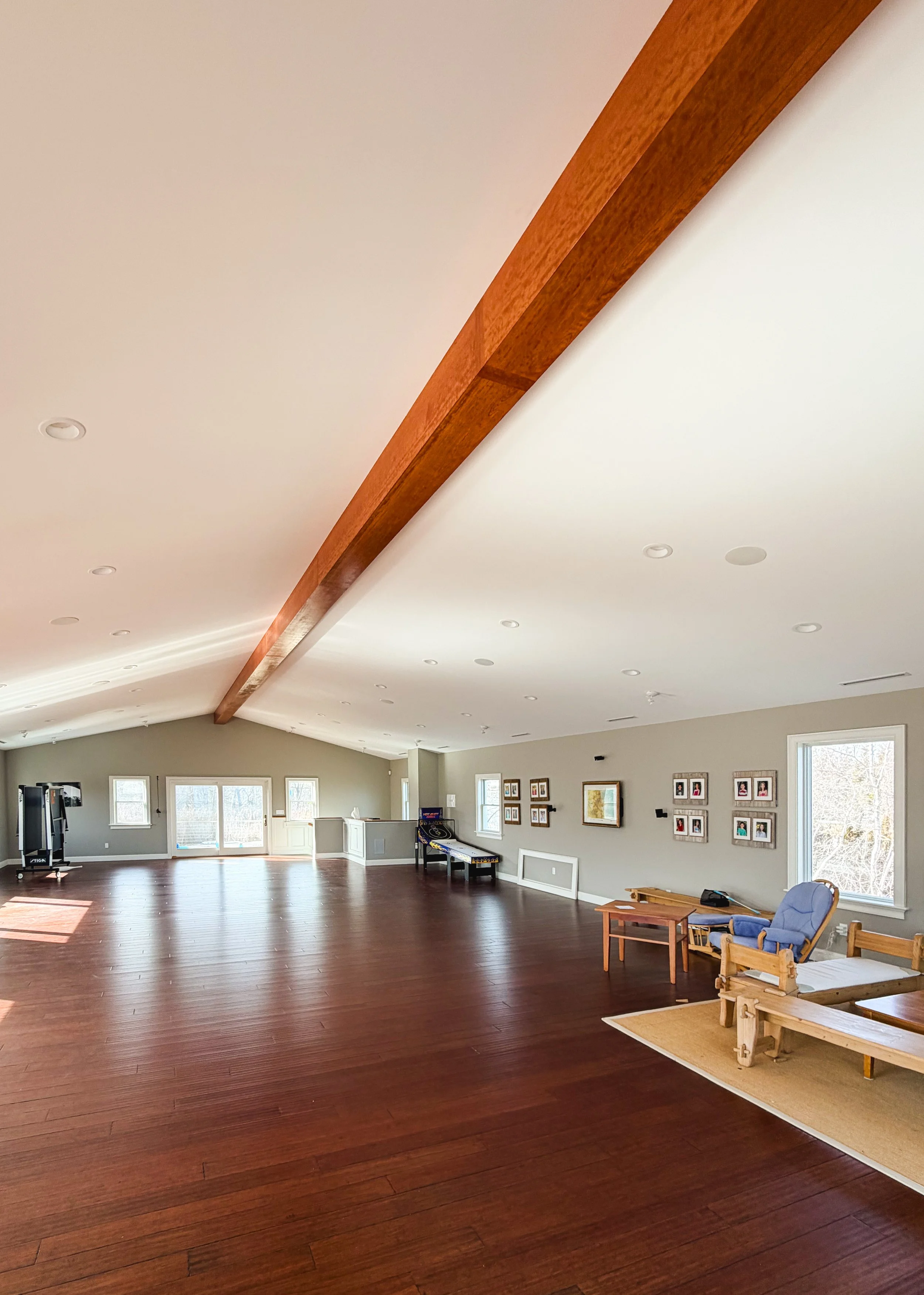 A spacious, bright living room with dark hardwood floors, multiple windows, a sloped ceiling with a wooden beam, and minimalist furniture and artwork on the walls.