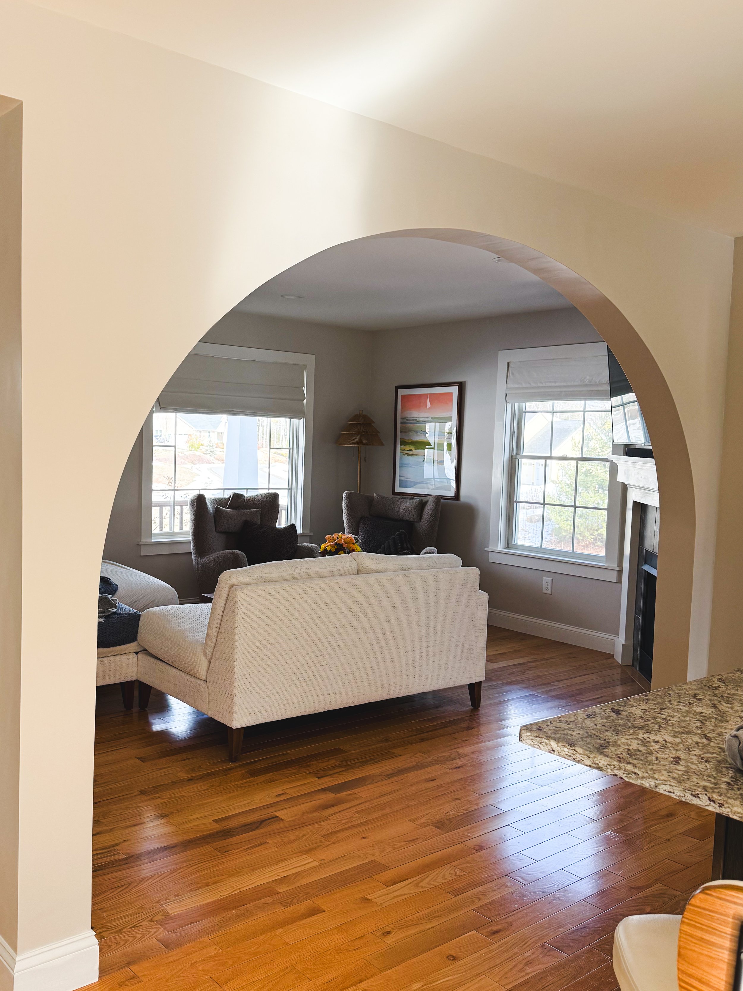 Living room with hardwood floors, a white sofa, two dark armchairs with pillows, large windows, artwork on the wall, and a fireplace, viewed through an arched doorway from the kitchen.