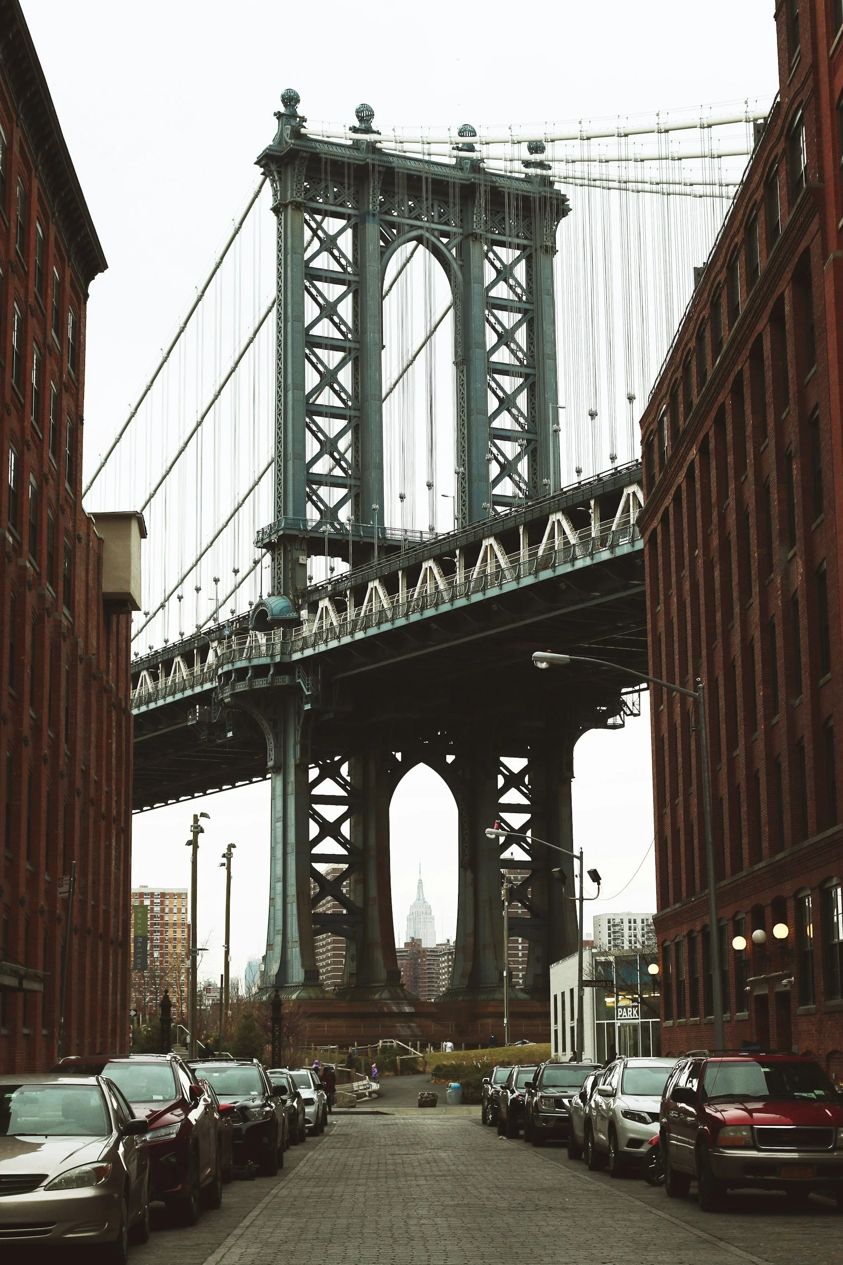View of the Brooklyn Bridge with Manhattan skyline, including the Empire State Building, in the background, seen from a parking lot surrounded by brick buildings