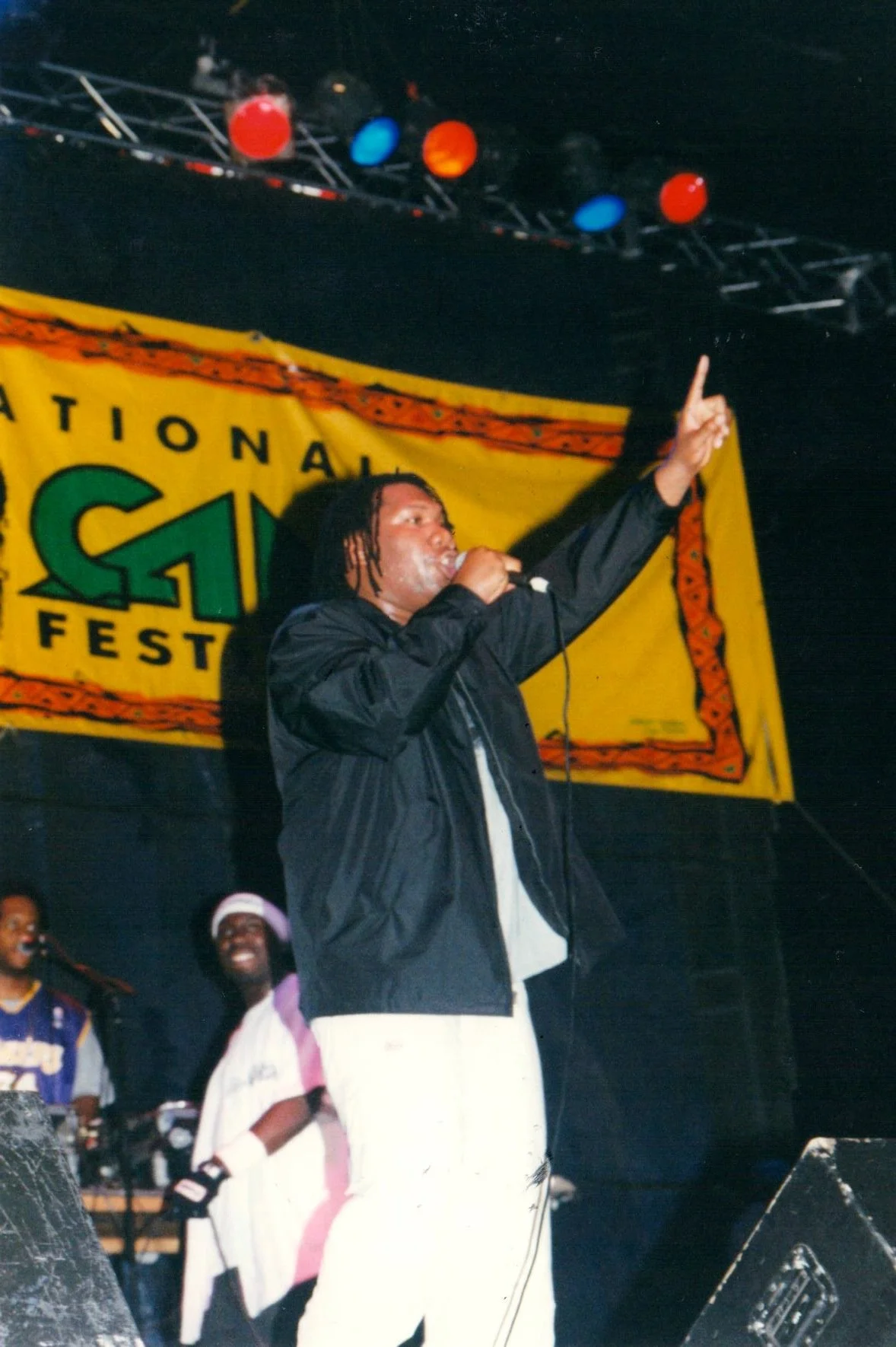 A performer with dreadlocks singing into a microphone on stage at an event with a yellow banner in the background that reads 'NATIONAL'. The performer is wearing a black jacket and light-colored pants, and two other people are visible in the background.