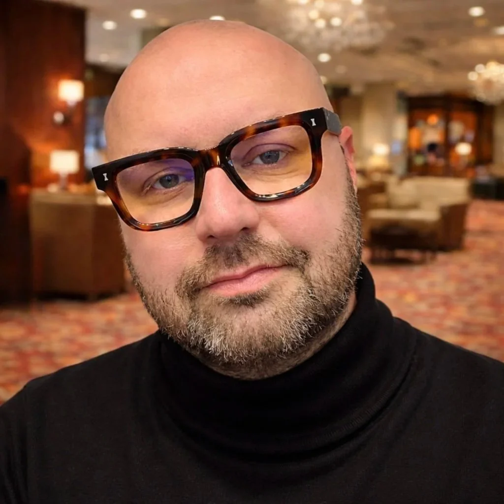 A bald man with a beard and wearing tortoiseshell glasses, dressed in a black turtleneck, in a warmly lit indoor setting with blurred furniture and decor in the background.