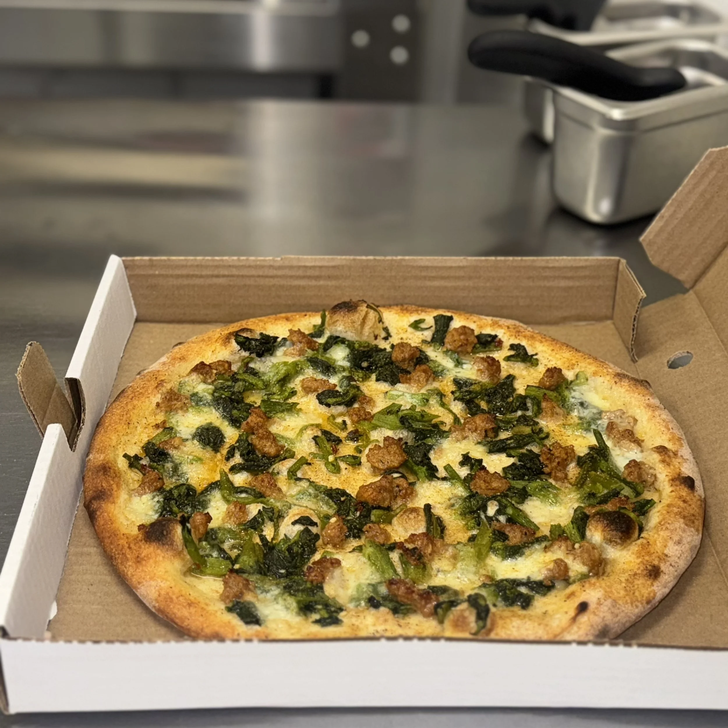 A pizza with green peppers, sausage, and cheese in an open cardboard box on a stainless steel countertop.