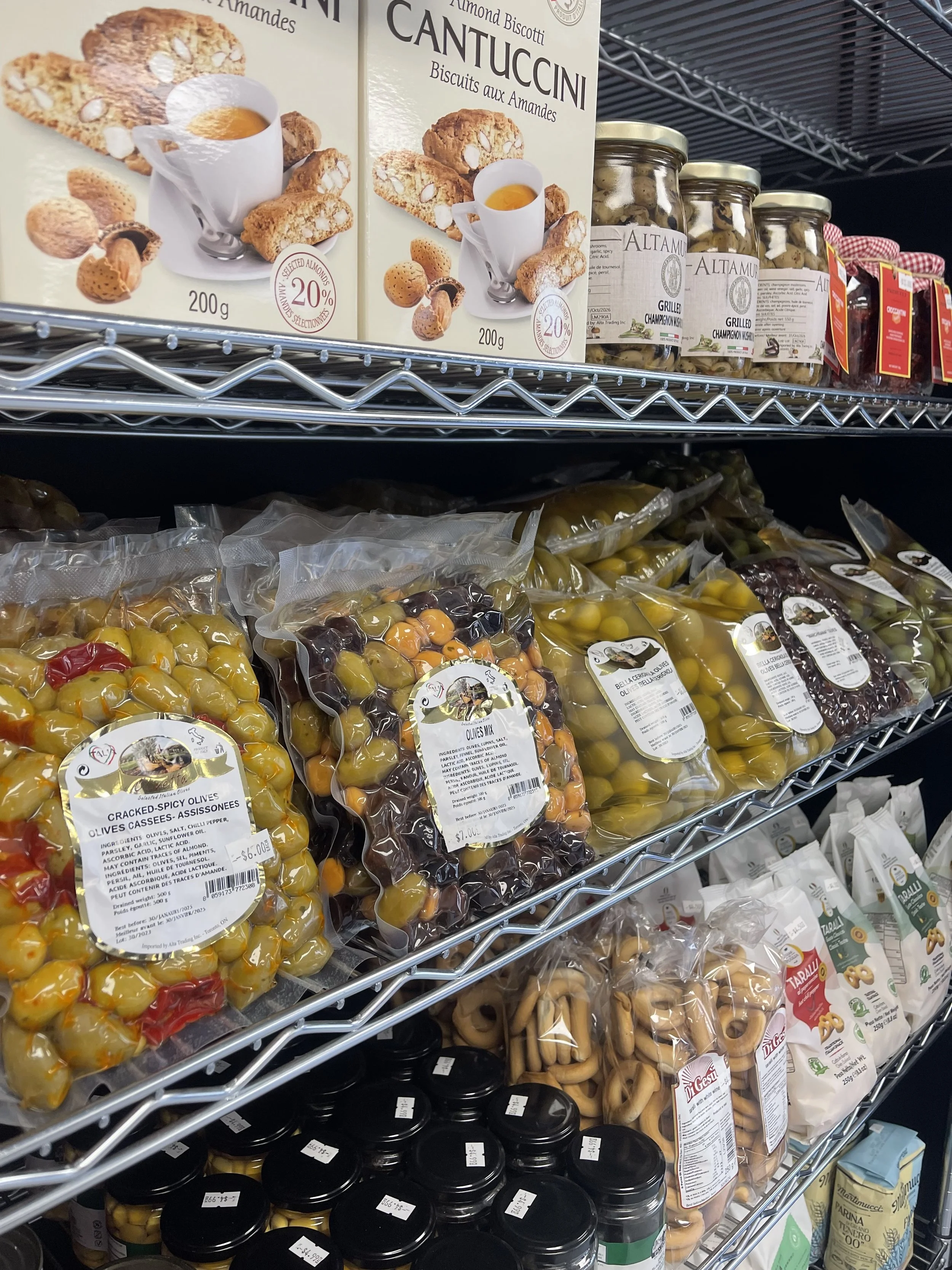 Shelf of packaged mixed olives, jars of grilled mushrooms, boxes of almond biscotti, and bags of baked goods in a grocery store.