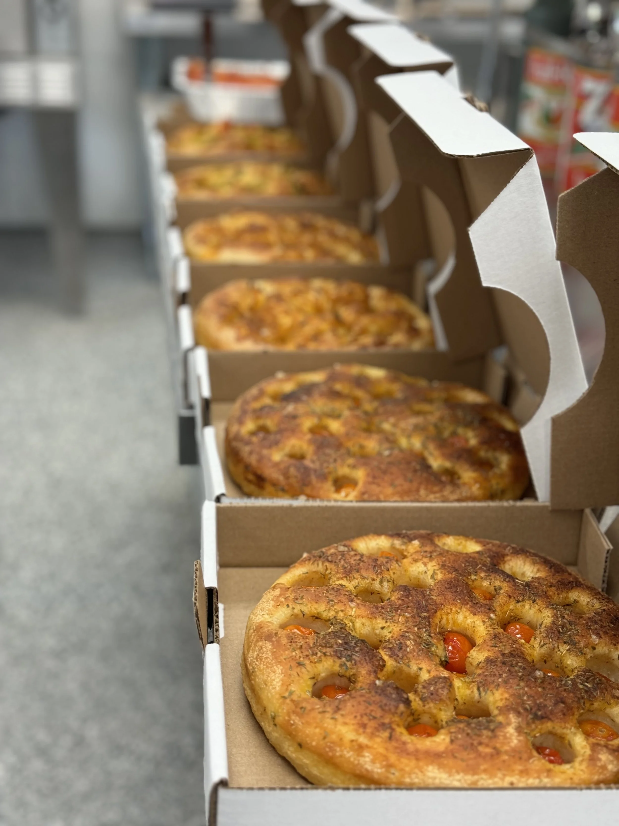 Line of six pizzas in cardboard boxes on a shelf, with one pepperoni pizza at the front and others varying in toppings further back, inside a store or pizzeria.