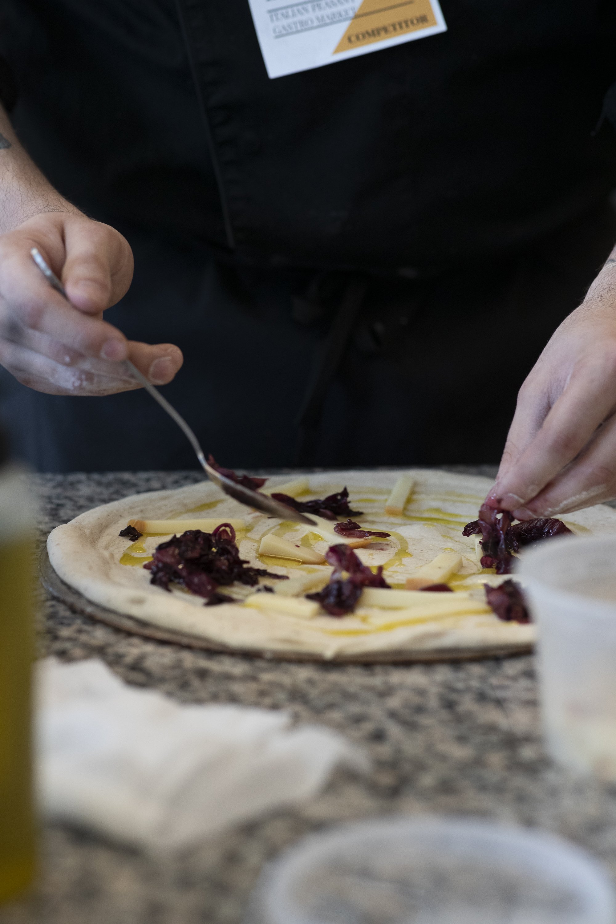 Person preparing a pizza with cheese, dried tomatoes, and olive oil on a granite countertop.