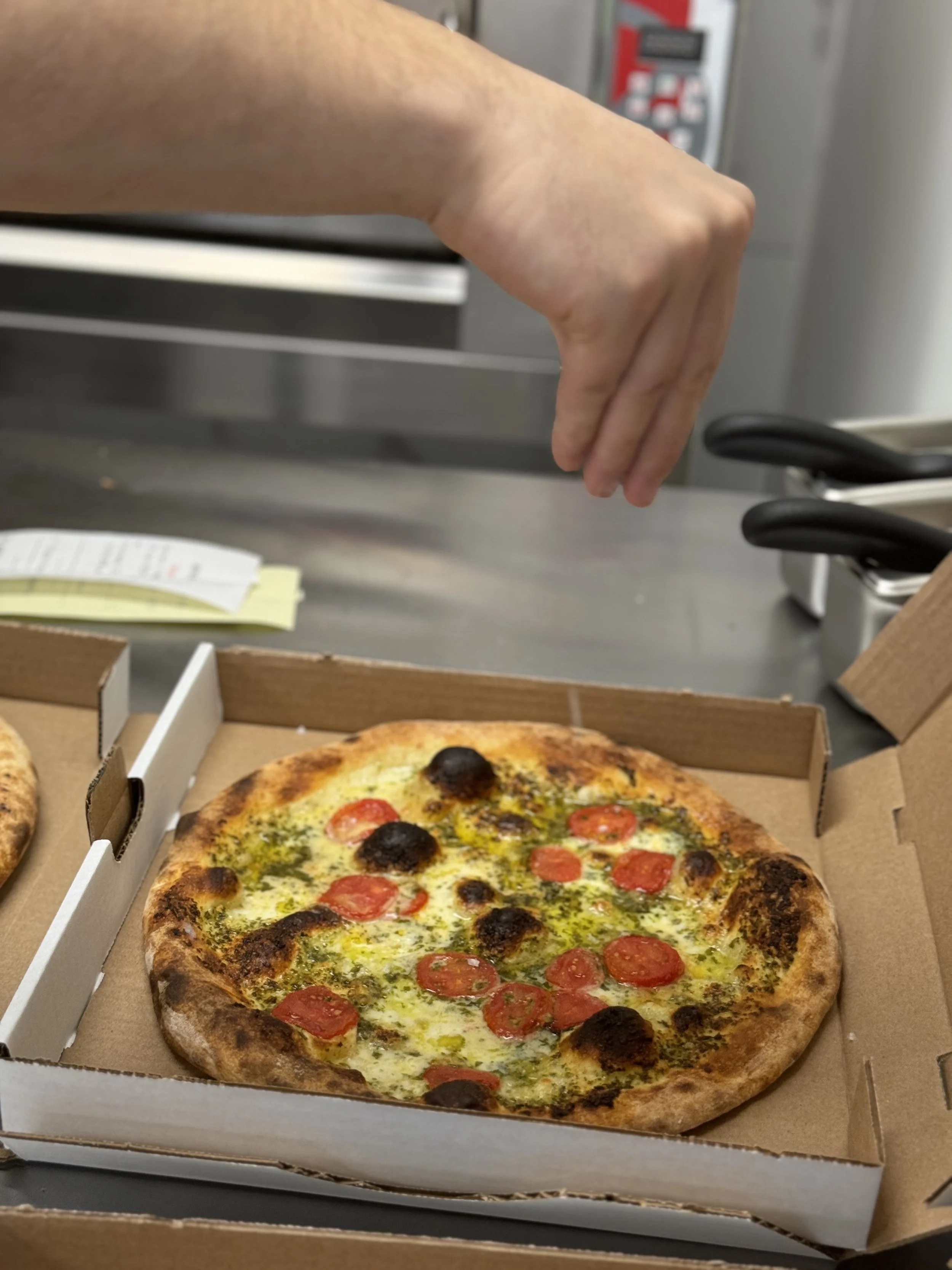 A pizza with cherry tomatoes, black olives, melted cheese, and green pesto in a cardboard takeout box with a hand about to grab the pizza.