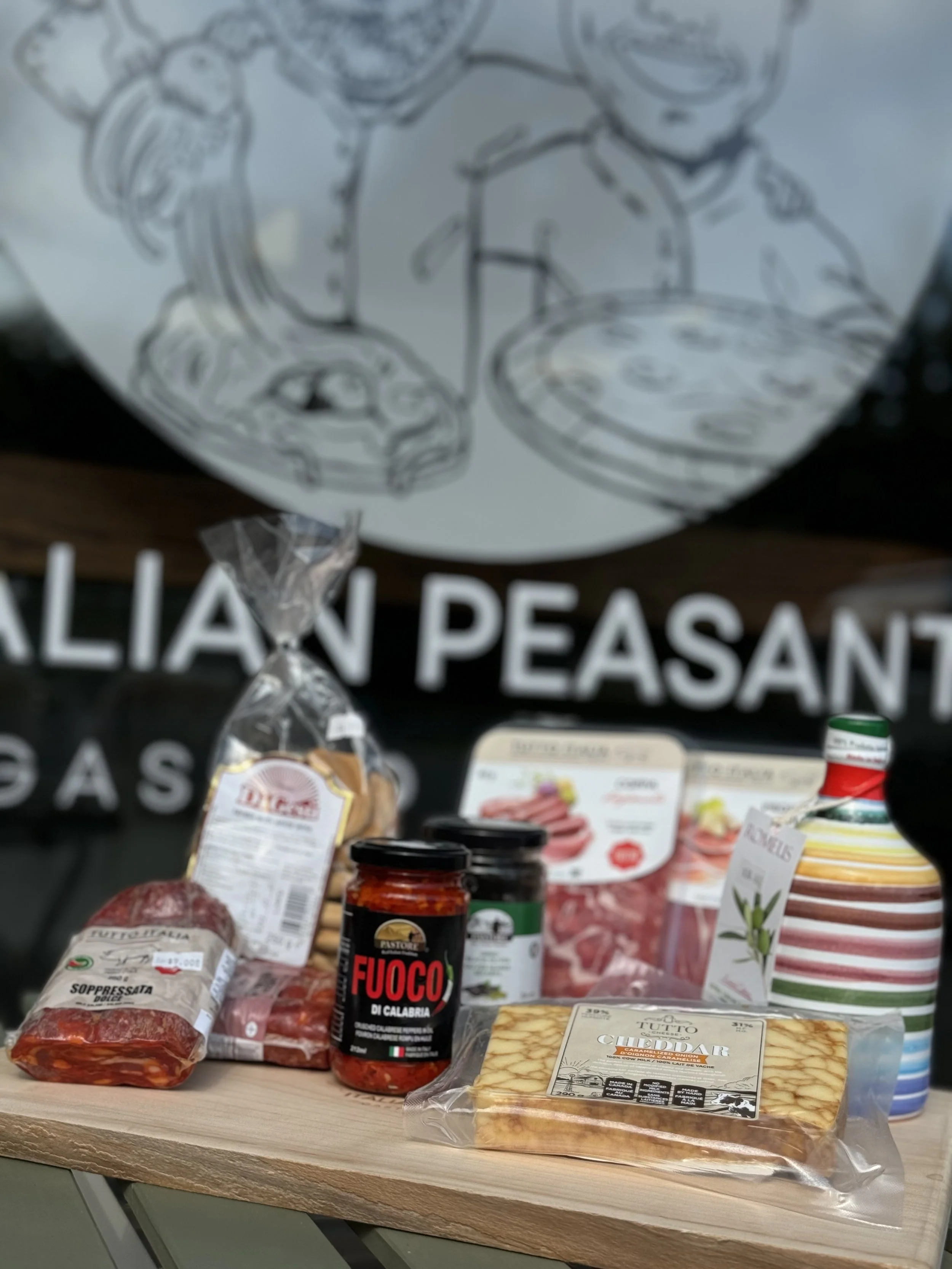 Assorted Italian food products on a wooden cutting board, including canned tomatoes, sliced cured meats, a grated cheese block, bottles of condiments, and sauce jars, with a sign reading 'ITALIAN PEASANT' in the background.