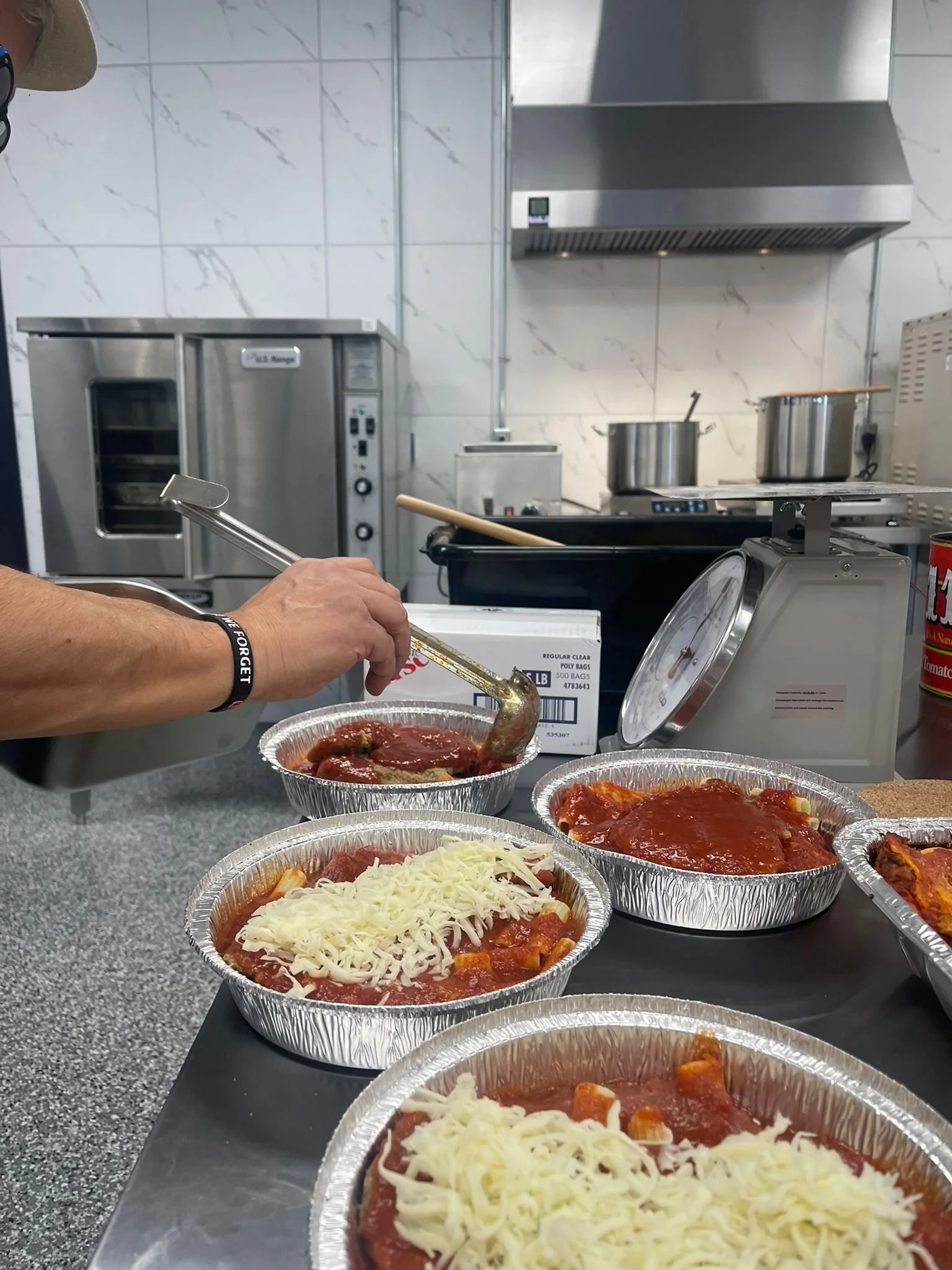 Aluminum trays filled with uncooked frozen lasagna and sauce on a kitchen counter, with a hand using a ladle to scoop sauce, in a commercial kitchen setting with ovens and stovetop in the background.