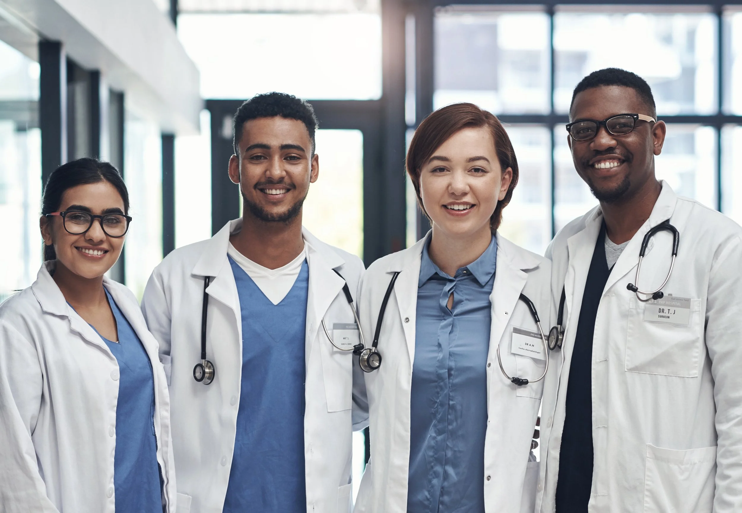 Four healthcare professionals in white coats stand together smiling at the camera, wearing stethoscopes in a bright hospital or clinic setting.