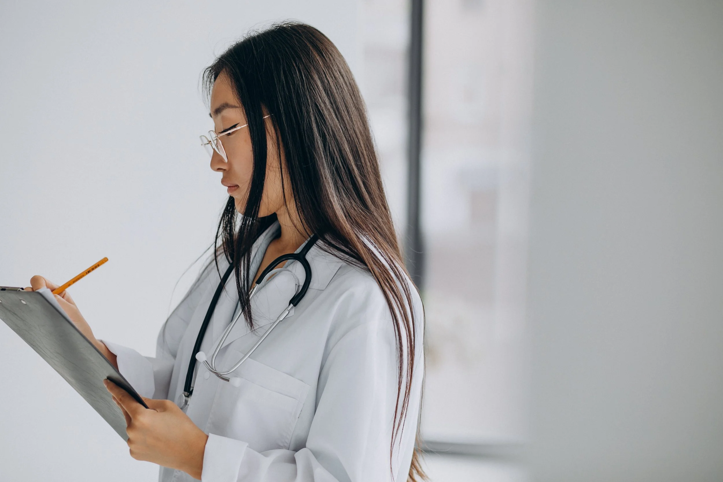 A doctor in a white coat with a stethoscope around her neck writes notes on a clipboard while standing in a bright clinical setting.