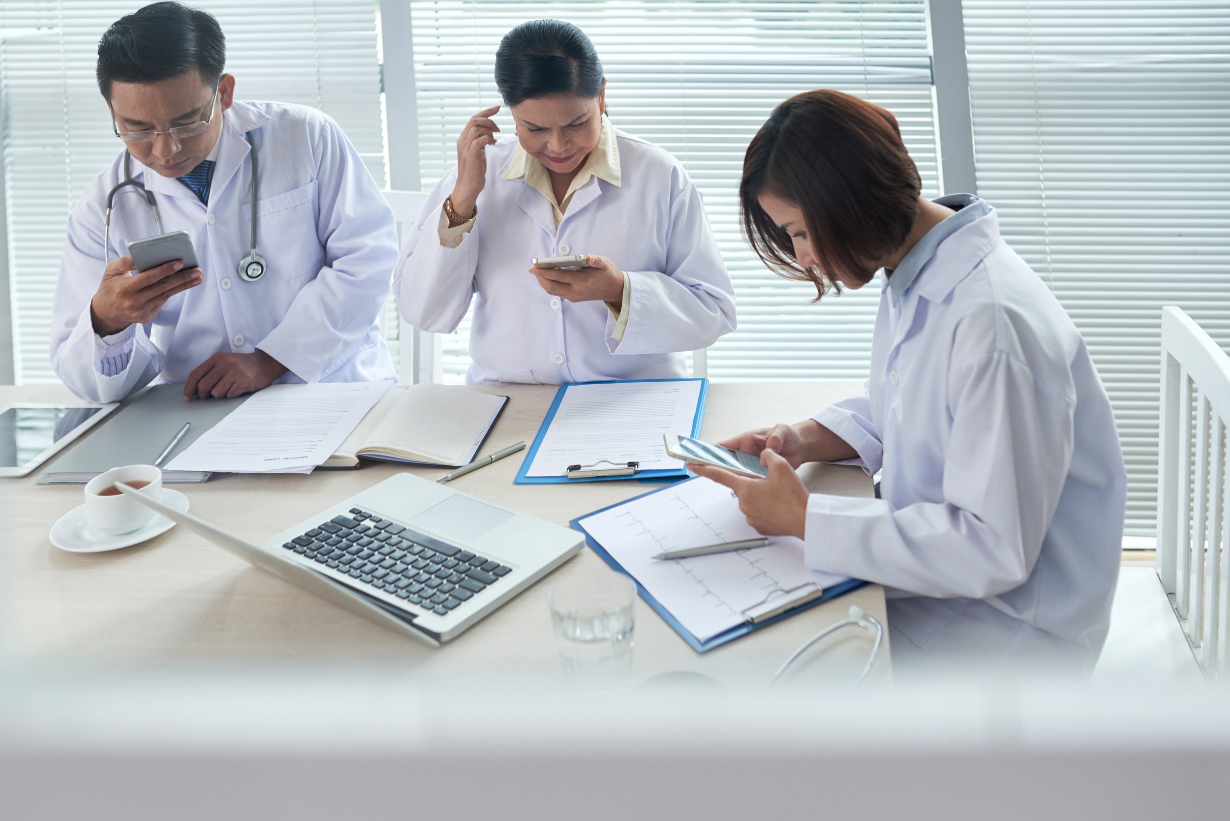 Three doctors in white coats sit at a table reviewing notes and charts while using smartphones, with a laptop and paperwork spread out in a bright office setting.