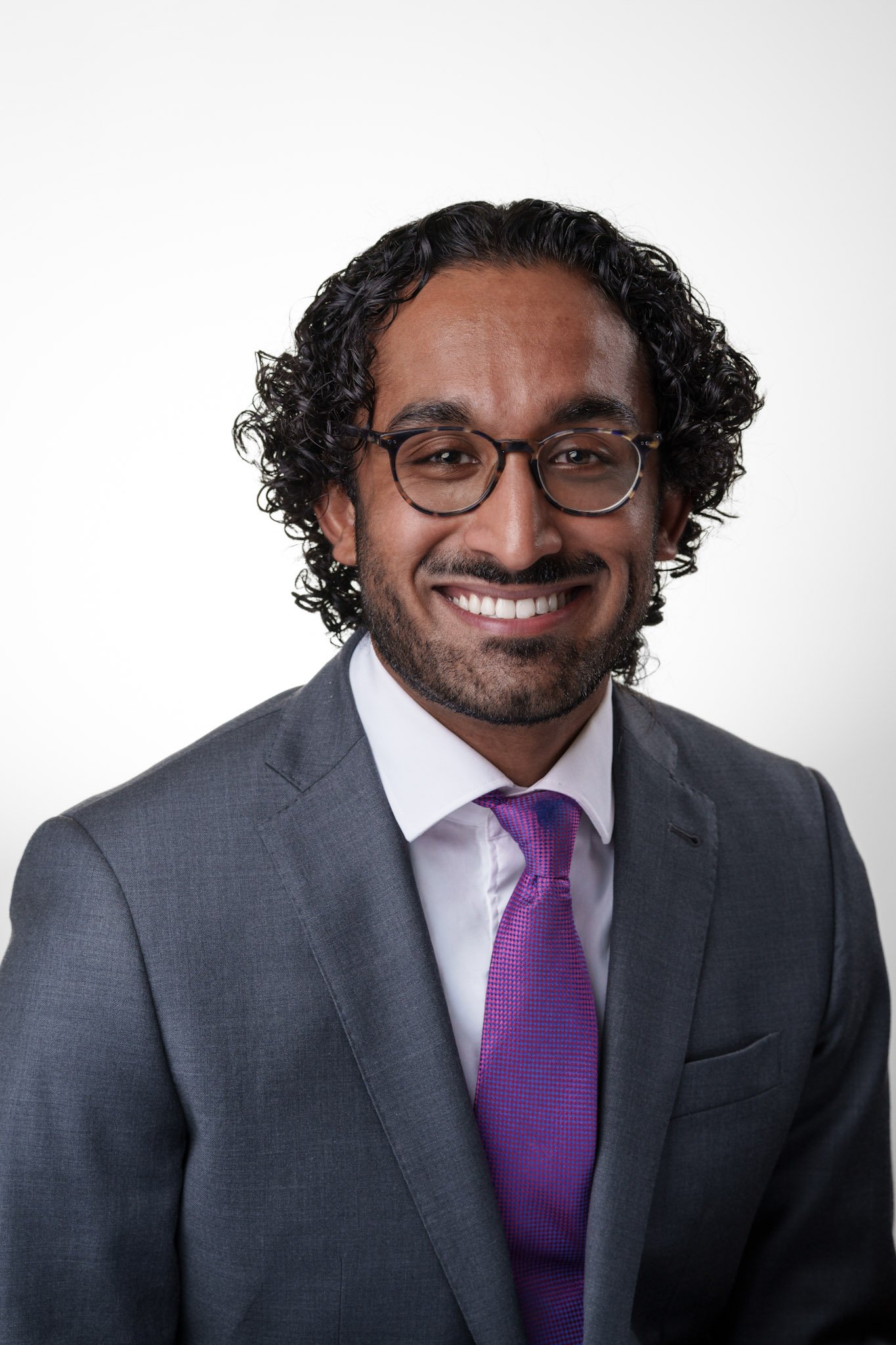 Man smiling in a professional headshot wearing glasses, a gray suit, and a pink tie against a light background.