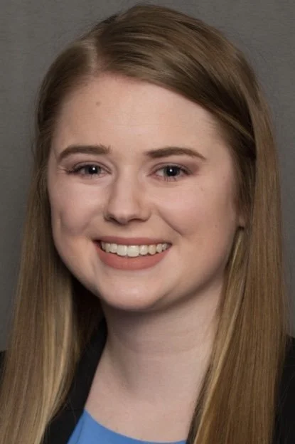 Woman smiling in a professional headshot with long hair, wearing a dark blazer and blue top against a neutral background.