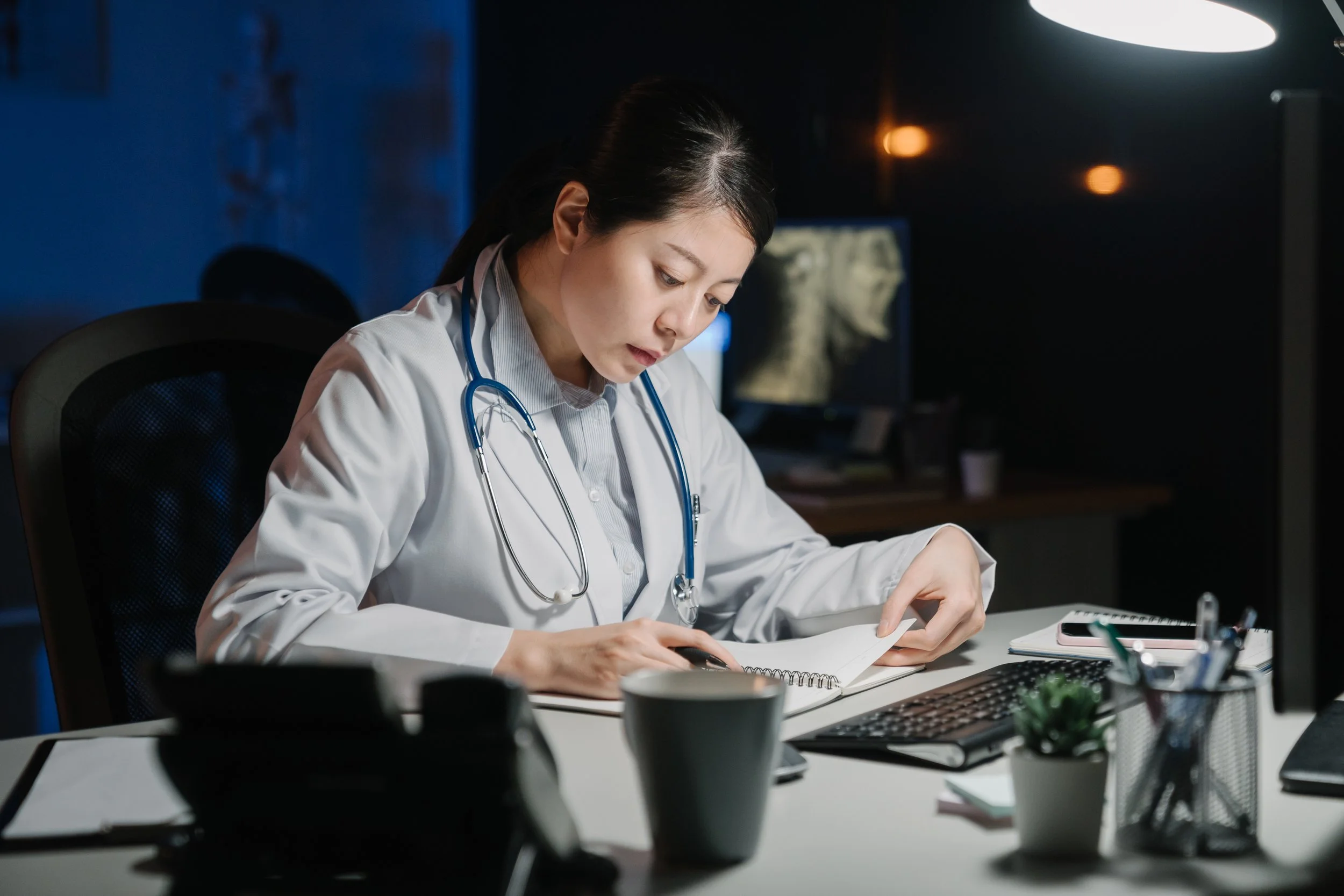 Female doctor reviewing medical notes at a desk late at night with a stethoscope around her neck.