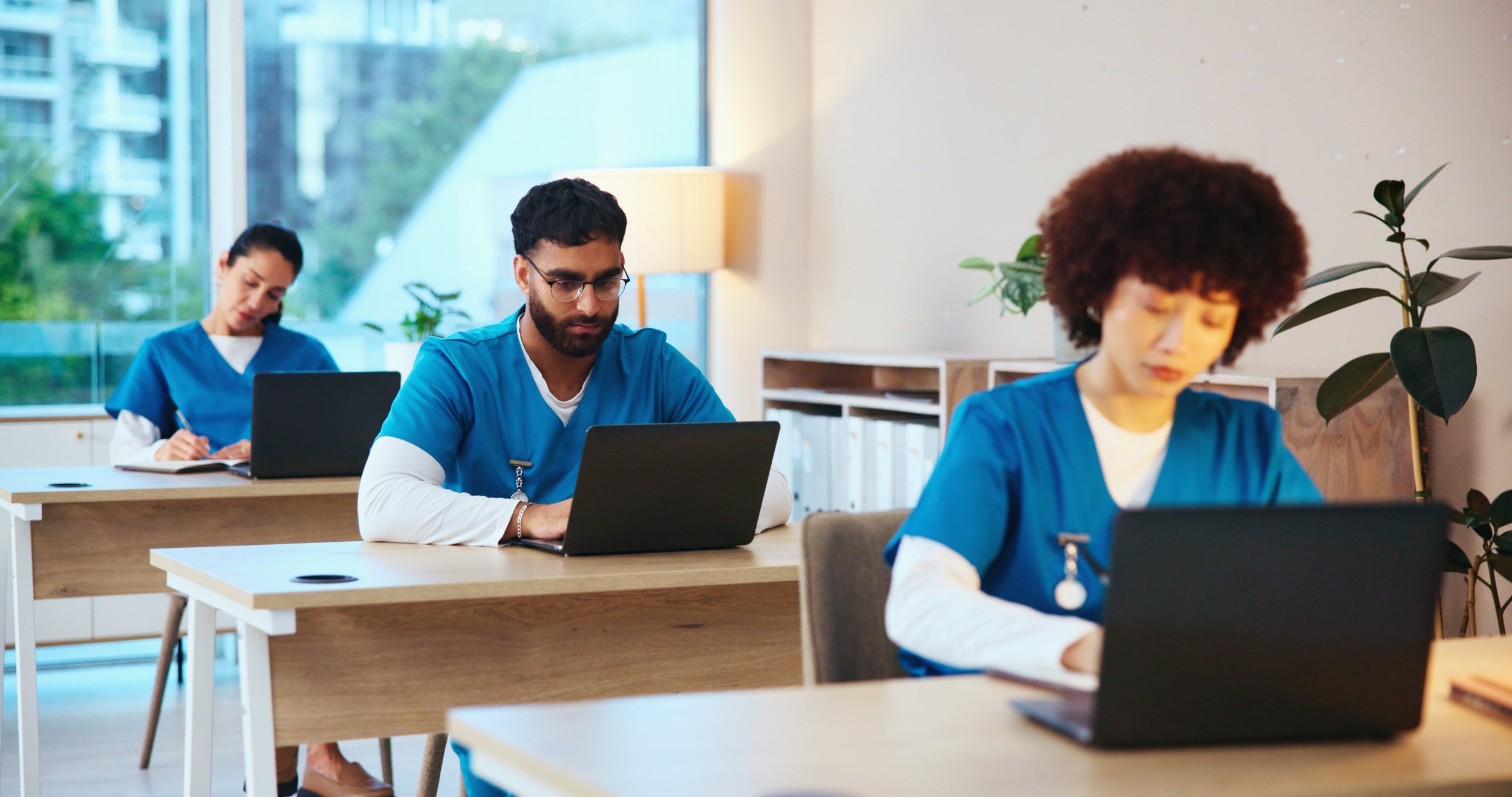 Three healthcare professionals in blue scrubs work independently at desks, using laptops and taking notes in a bright medical study or training environment.