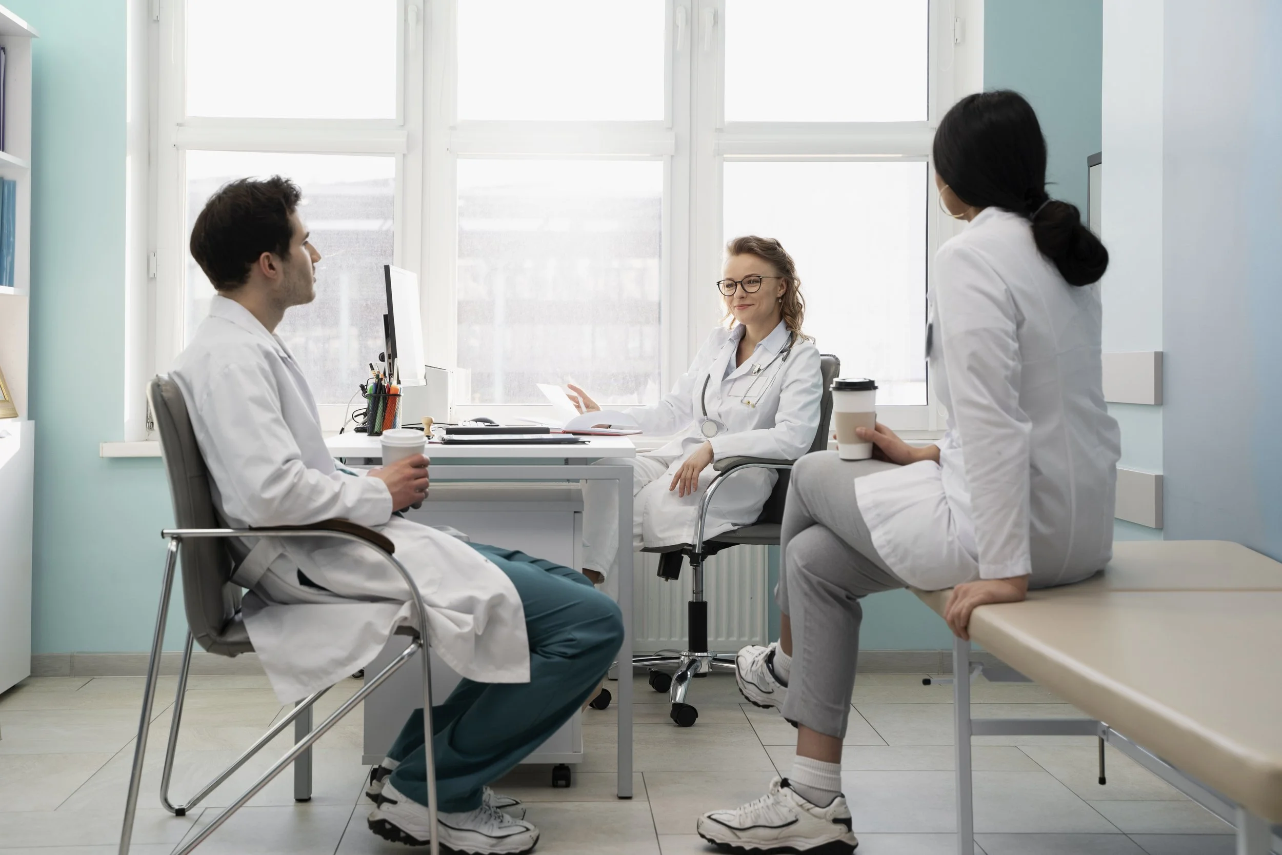 Three doctors in white coats sit together in a bright medical office, discussing patient care while holding coffee cups during a team meeting.