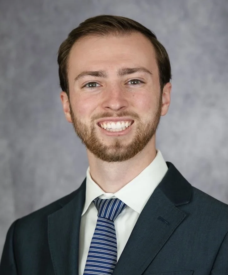 Man smiling in a professional headshot wearing a dark suit and striped tie against a gray background.