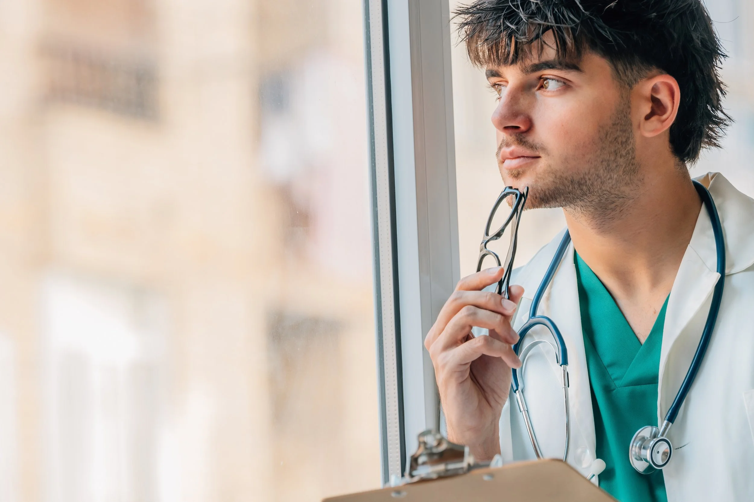 Thoughtful medical student in a white coat and scrubs holding glasses while looking out a window.