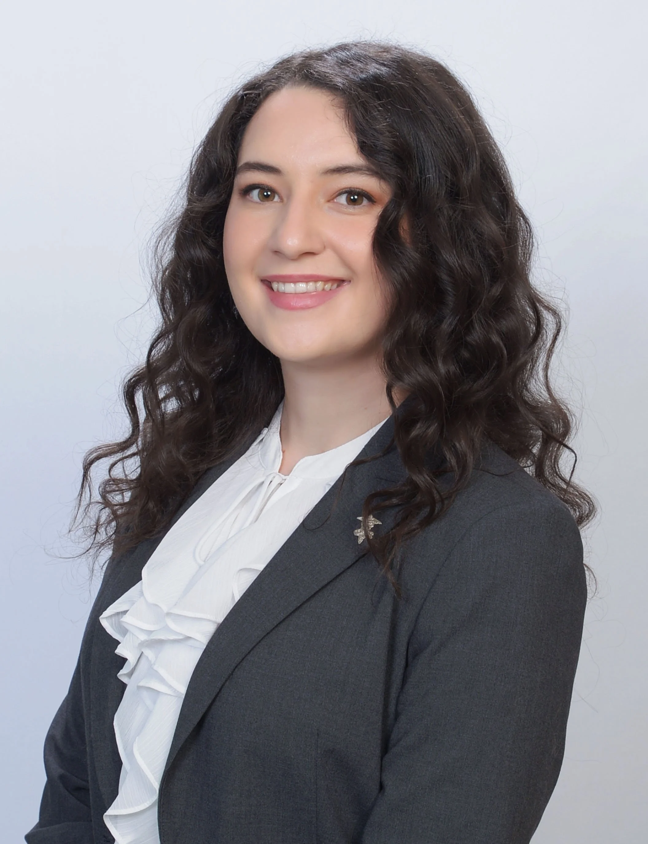 Professional headshot of a woman with long curly hair wearing a dark blazer and white blouse against a light background.