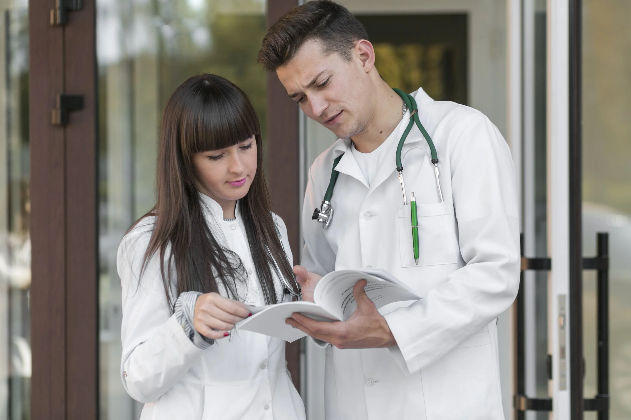 Two medical professionals in white coats stand together reviewing a medical textbook, with one pointing to a page while discussing the content outside a healthcare facility.