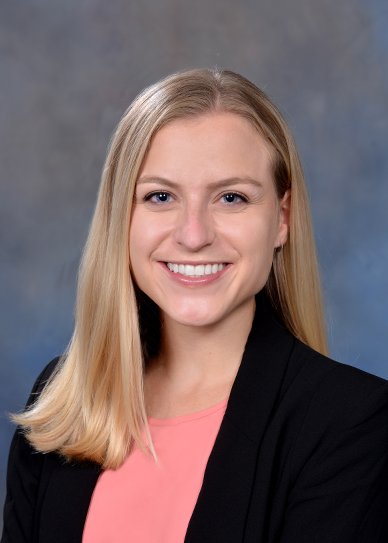 Woman smiling in a professional headshot wearing a black blazer and coral top against a neutral studio background.