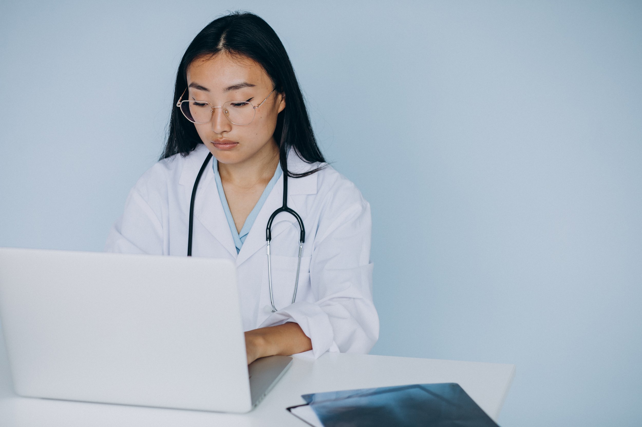 A doctor wearing glasses and a white coat works on a laptop, reviewing medical images at a desk in a clean, minimal clinical setting.