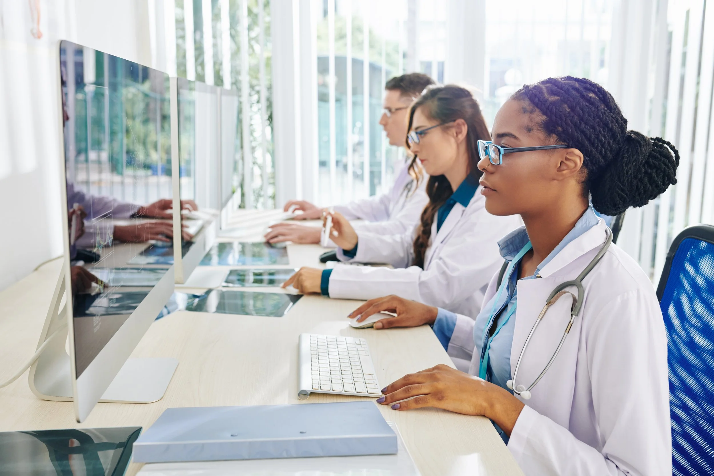 Three healthcare professionals in white coats work at desktop computers, reviewing medical images and data in a bright clinical office.