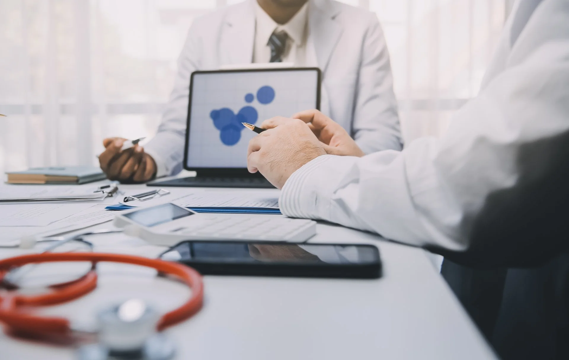 Two medical professionals in white coats sit at a desk discussing data, with one pointing at a tablet displaying blue circular charts, surrounded by a stethoscope, documents, and a smartphone.