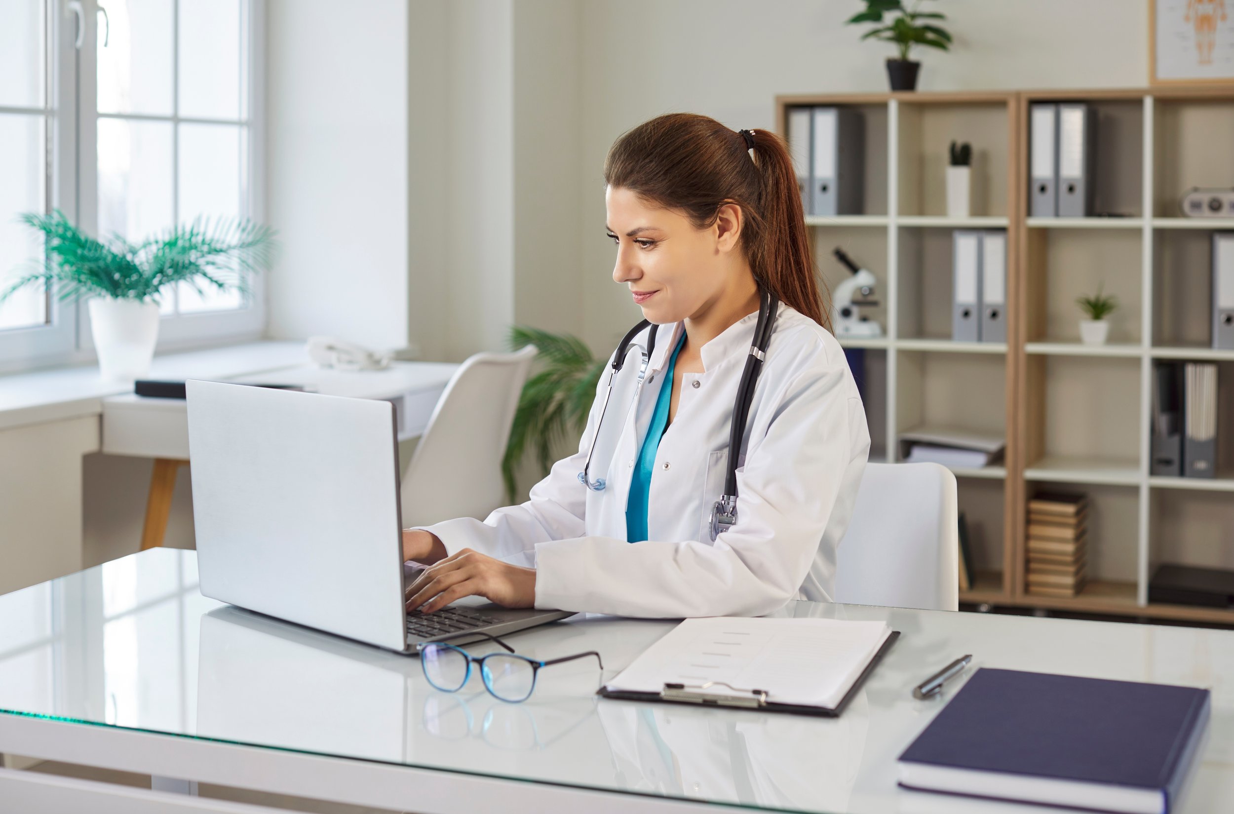 Female doctor in a white coat working on a laptop at a desk in a modern medical office.